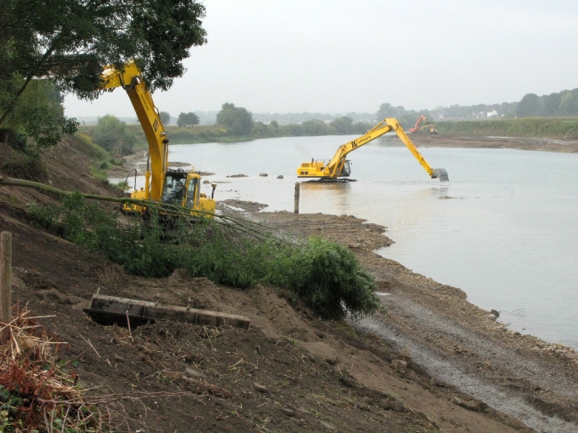 Ter hoogte van het huis van Bours worden nog wat bomen weggehaald. Hier wordt de oever beschermd met een laag beton omdat de ondergrond te instabiel is. Op de achtergrond staat de graafmachine zelfs tot in het midden van de Maas om kiezel te winnen voor de aanleg van de werkweg. Ter hoogte van het huis van Bours worden nog wat bomen weggehaald. Hier wordt de oever beschermd met een laag beton omdat de ondergrond te instabiel is. Op de achtergrond staat de graafmachine zelfs tot in het midden van de Maas om kiezel te winnen voor de aanleg van de werkweg.