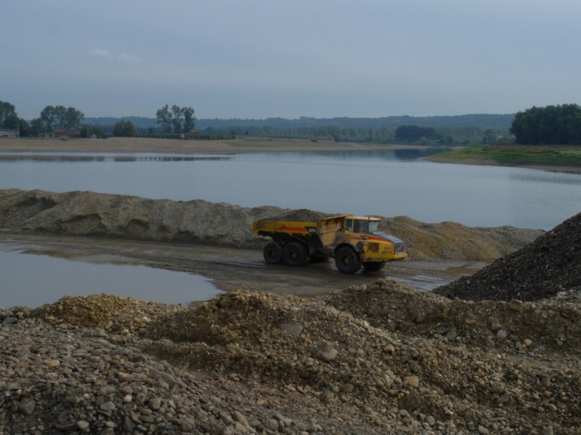 Dumpers vervoeren de afgegraven dekgrond naar de dekgrondberging. De waterplas op de achtergrond geeft het beeld weer dat ons waarschijnlijk te wachten staat. De graafmachines hebben hier al tot op een diepte van 36m boven NAP de kiezel weggegraven. 