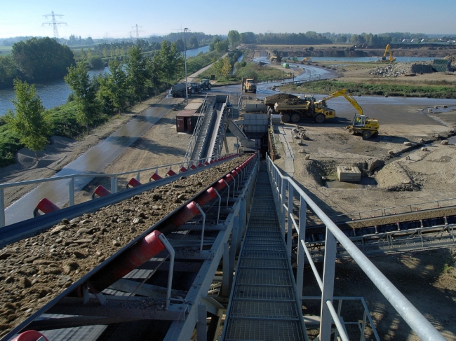 Bovenop de installatie staande, kijkend naar het zuiden, zie je links het Julianakanaal en aan de voet van de installatie zie je een dumper toutvenant lossen. Verder naar achter de dekgrondberging in het Hartelsteinerveld die alweer gedeeltelijk opgevuld is. 